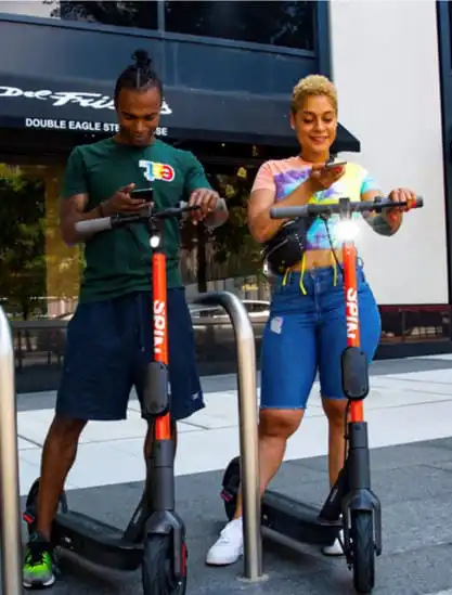Two people riding electric scooters on city sidewalk